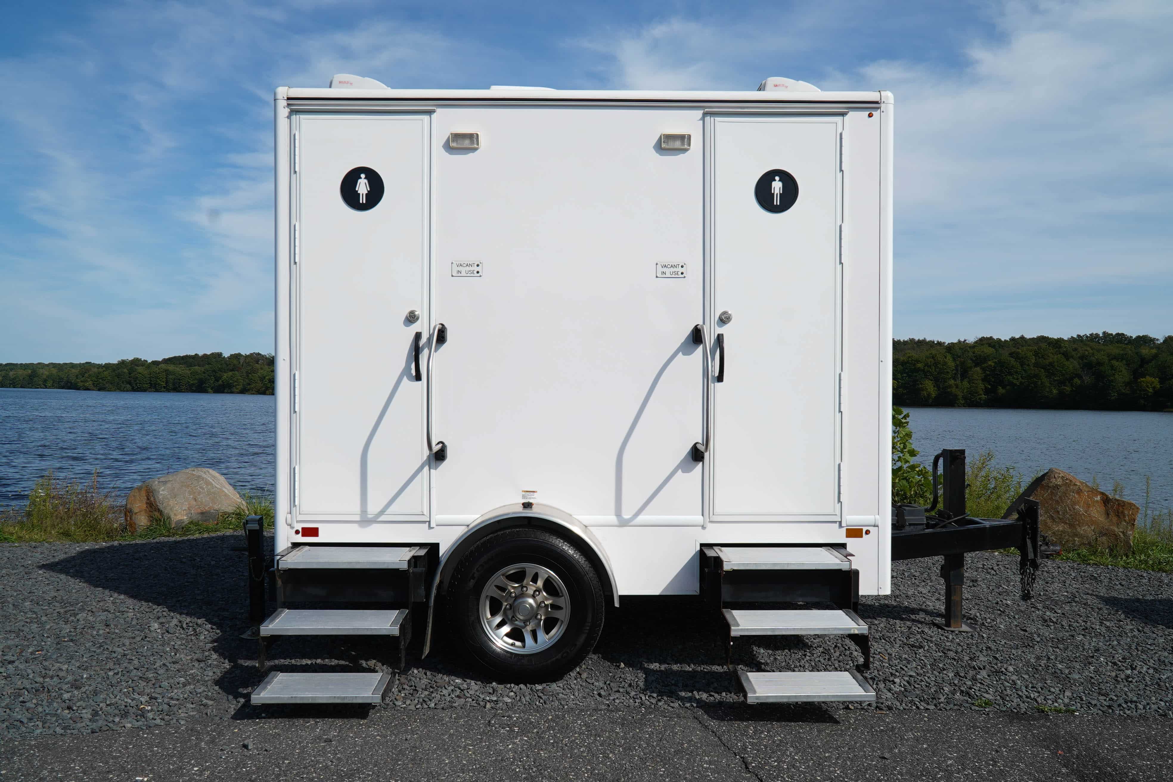 A portable restroom trailer is positioned in front of a tranquil lake. It features two entrances with gender symbols, one for women and one for men, and has signs indicating the availability of each stall. The exterior is white, and there are steps leading up to each door. In the background, lush greenery surrounds the water.