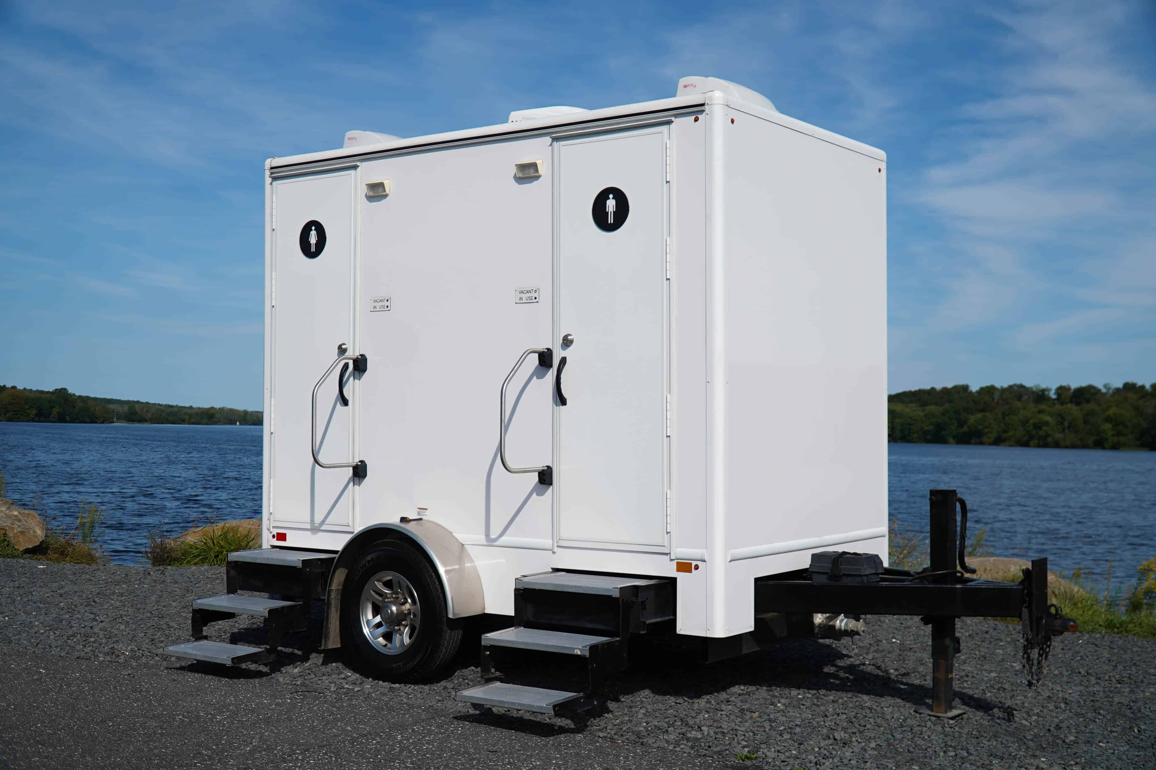 A white portable restroom trailer with two doors, located next to a body of water. Steps lead up to each door, and the surrounding area features gravel and greenery under a clear blue sky.