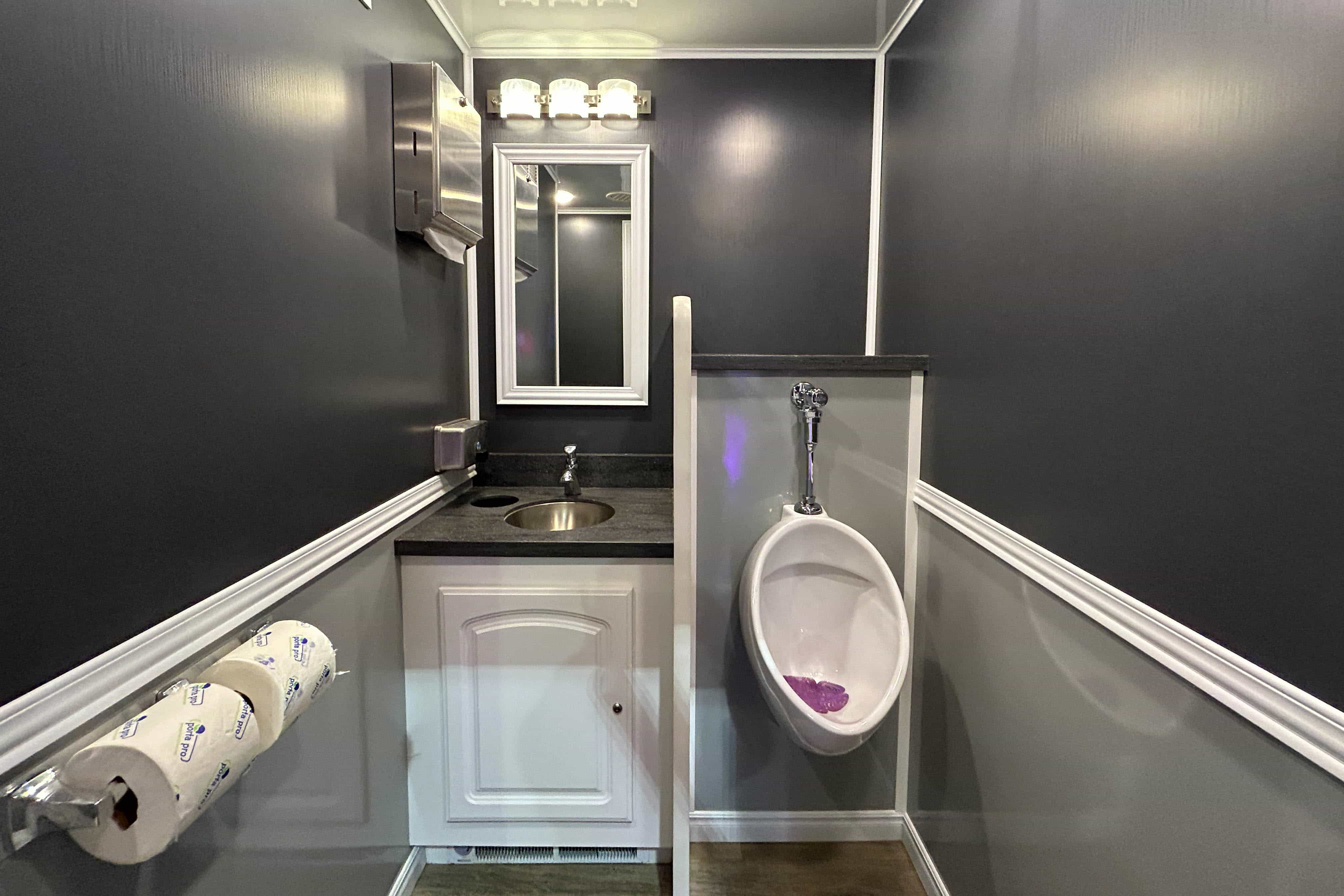 A clean and modern bathroom interior featuring dark gray walls, a small sink with a countertop, a mirror above it, and a white urinal on the right side. There is a roll of paper towels mounted on the wall, and a silver hand dryer is also visible. The space is well-lit with overhead lighting.
