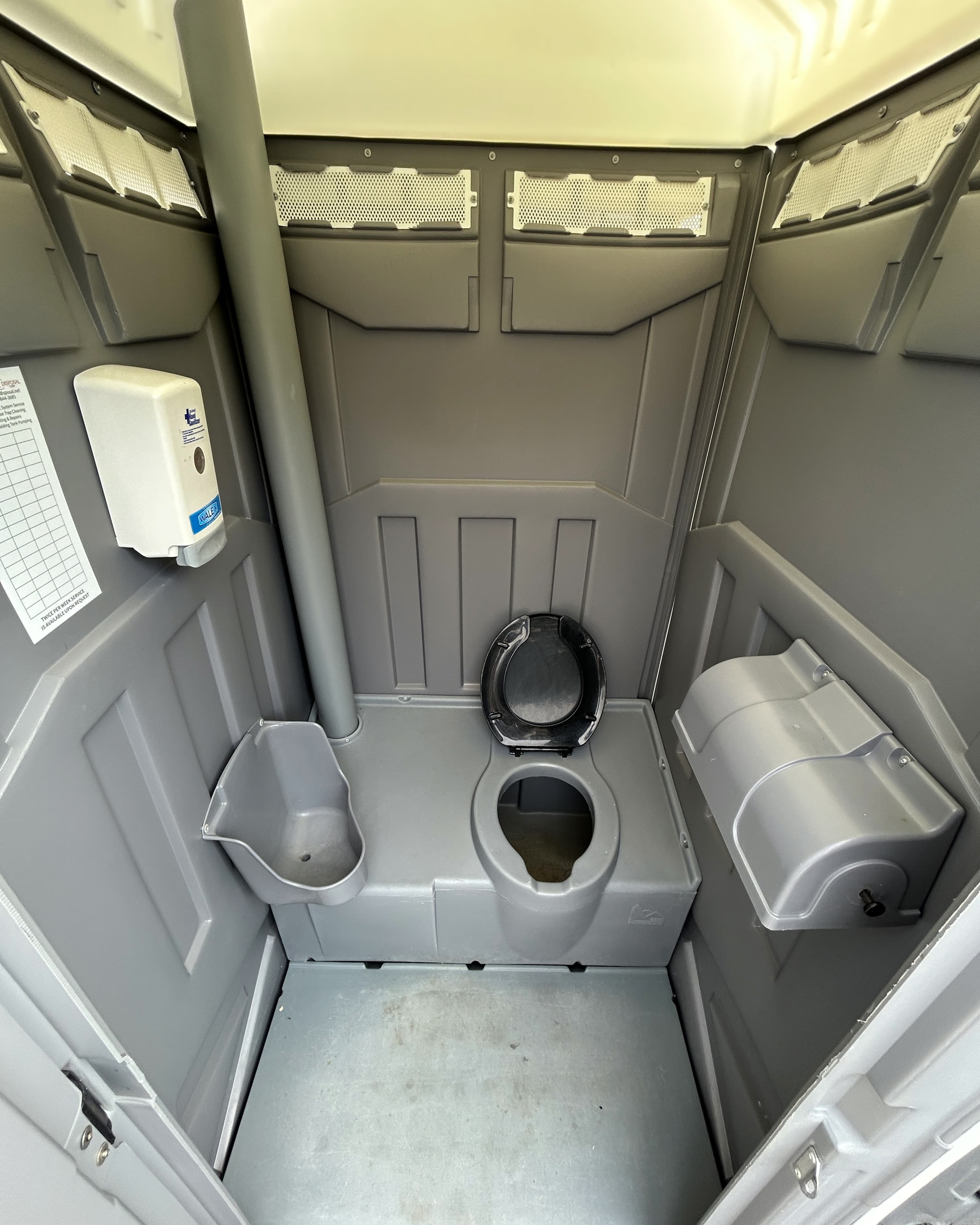 A clean, gray portable toilet interior. The space includes a toilet seat, a hand sanitizer dispenser on the wall, and a small shelf for personal items. The walls are made of a durable material, and there are vents near the top for air circulation. The floor is flat and also gray.