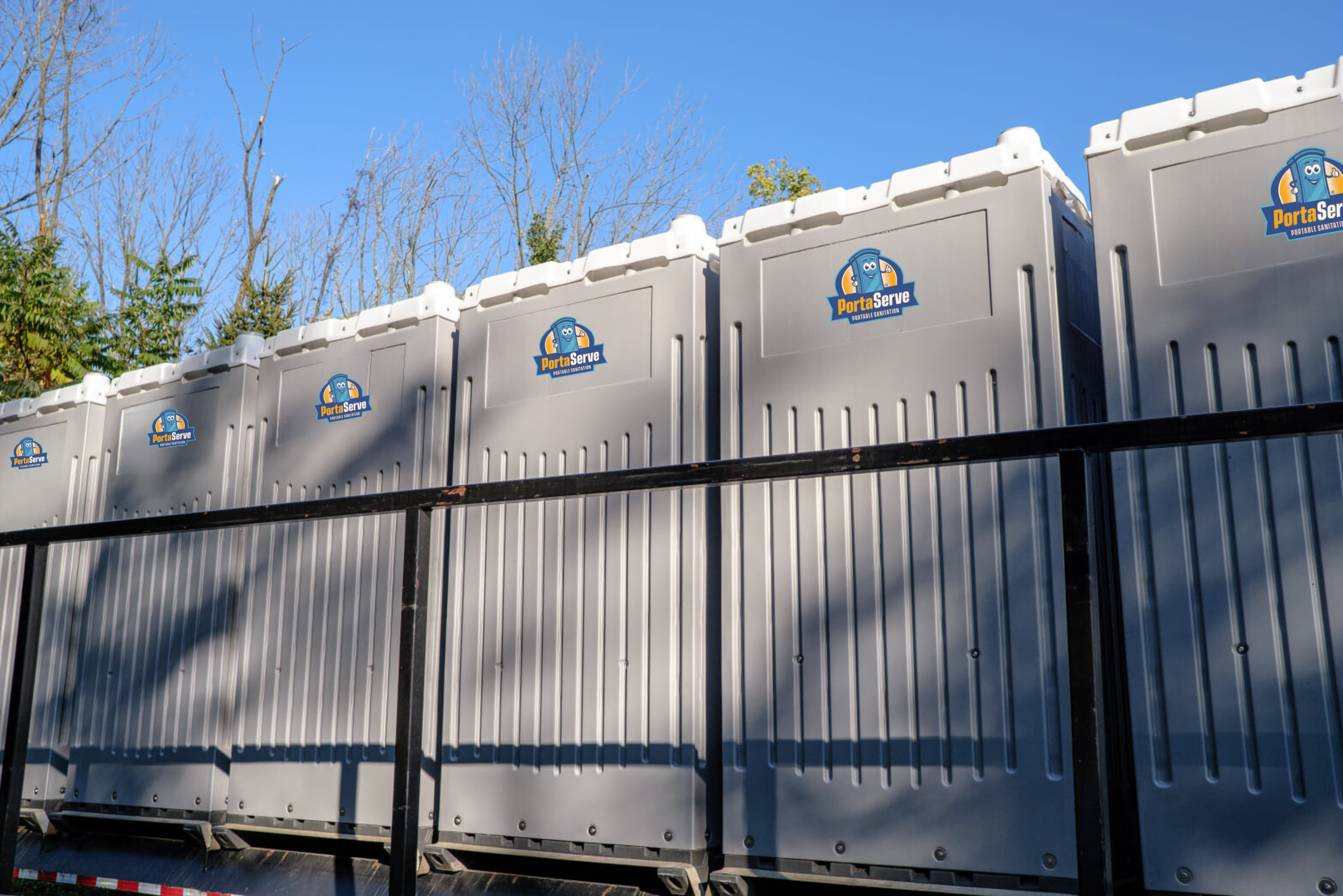 A row of gray portable toilets with the PortaServe logo, positioned on a truck against a clear blue sky and bare trees in the background.
